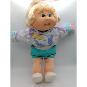 May include: A doll with blonde yarn hair, blue eyes, and a single visible tooth. The doll wears a long-sleeved shirt featuring a white, purple, yellow, and teal geometric pattern, paired with solid teal shorts and plain white shoes.