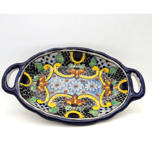 May include: Oval-shaped, hand-painted ceramic serving dish with handles. The dish features a dark blue rim and handles, with a central design of flowers and leaves in blue, yellow, and brown against a black background.