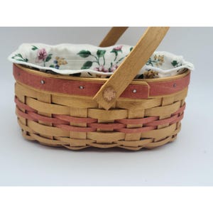May include: A woven basket with a wooden handle and a floral patterned lining. The basket features a light brown base with red accents and a red band around the top. The interior lining is white with a floral pattern.