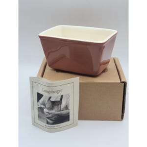 May include: A Longaberger brand, terracotta-colored ceramic butter dish with a white interior. The dish is sitting on a brown cardboard box with a Longaberger label.