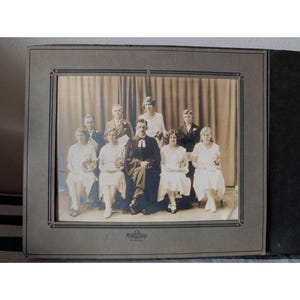 May include: Sepia-toned photograph of a group of people, likely from the early 20th century. The subjects are formally dressed, seated in front of a curtain backdrop. The photo is framed in a dark gray matte.