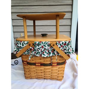 May include: A light brown woven picnic basket with a wooden top and handles. The top has a small table supported by four wooden legs. The basket is decorated with a fabric trim featuring a green and red berry pattern.
