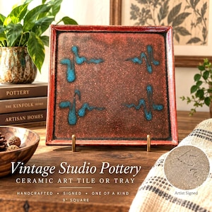 May include: A square ceramic tray with a textured, deep red and brown speckled glaze. The surface features four distinct, raised turquoise blue abstract markings arranged in a symmetrical pattern. The tray is displayed on a gold metal stand.