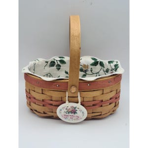 May include: Woven basket with a wooden handle and a floral patterned lining. The basket has a pink band around the top and a decorative tag that reads "Happy Mother's Day 1999" with a floral design.