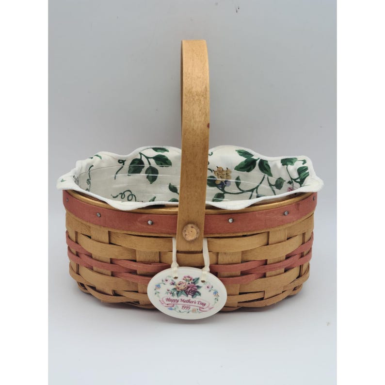 May include: Woven basket with a wooden handle and a floral patterned lining. The basket has a pink band around the top and a decorative tag that reads "Happy Mother's Day 1999" with a floral design.