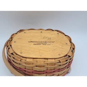 May include: A woven basket with a light brown wooden base and rim, featuring a red-toned band around the middle. The basket has a handle and the text "Longaberger Baskets Handwoven Dresden, Ohio USA" is visible on the base.