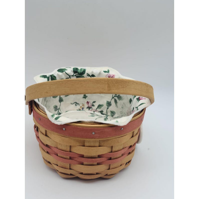 May include: A woven basket with a wooden handle and a pink band around the middle. The basket's interior is lined with a floral fabric featuring pink roses and green leaves. The basket is designed for carrying items.