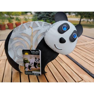 May include: A solar-charged panda sculpture featuring a white face with black eye patches, blue gemstone eyes, and a black nose. The body has a silver-toned, intricate cutout pattern. A product tag labelled Garden Meadow LED Lighted Outdoor Sculpture is attached.