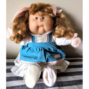 May include: A doll with curly brown hair tied with pink ribbons, featuring dark eyes and a round face. The doll wears a white long-sleeved blouse under a teal blue pinafore dress with a floral patterned bodice, white ruffled trim, and white shoes.