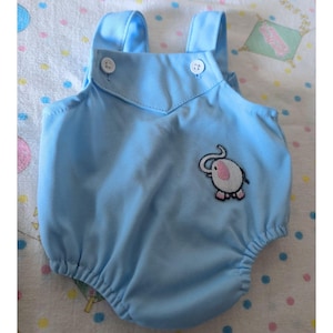 May include: A light blue sleeveless romper featuring a V-shaped front bib with two white buttons at the top straps. The garment includes an embroidered patch of a white elephant with pink ears and feet positioned on the lower right side of the front.