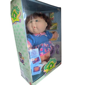 May include: A Cabbage Patch Kids Fun to Feed Baby doll with brown hair and green eyes, wearing a blue dress with a pink pattern. The doll is inside a box with accessories including a purple spoon, a bowl, and play food boxes labeled Mott's and Peas & Carrots. The box features text explaining the doll's interactive features.