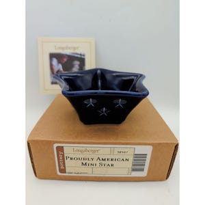 May include: A navy blue, star-shaped mini dish with three white star cutouts on the front. The dish sits on a brown cardboard box with the text "Proudly American Mini Star" and the Longaberger Pottery logo.