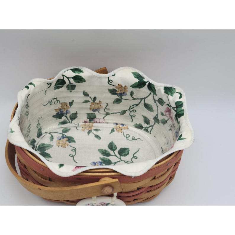 May include: A woven basket with a red-brown rim and a wooden handle. The interior features a floral patterned fabric lining with green leaves and yellow and pink flowers. The basket has a decorative wooden clasp.