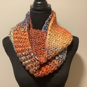 May include: A hand-knitted infinity scarf in a blend of orange, brown, blue, and yellow yarns. The chunky knit scarf is displayed on a black mannequin. The color palette creates a warm, autumnal feel, perfect for cold weather.