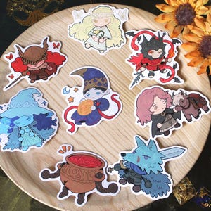 May include: A collection of colorful character stickers on a wooden plate. The stickers feature various fantasy-themed illustrations, including a witch, a knight, and a wolf with a sword. Decorative sunflowers and dice are in the background.