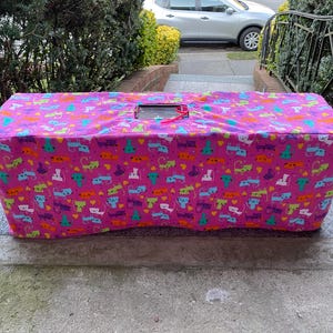 Op de afbeelding: Een rechthoekige, roze stoffen hoes met een herhaald patroon van kleurrijke cartoonkatten. De hoes heeft een rechthoekige opening aan de bovenkant met een rood lint. Hij ligt op een stenen oppervlak.