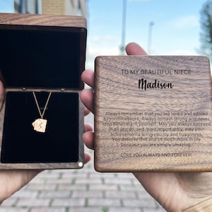 Może przedstawiać: Drewniane pudełko z złotym naszyjnikiem w środku. Pudełko ma wygrawerowany napis: "Do mojej pięknej siostrzenicy Madison. Pamiętaj zawsze, że jesteś kochana i uwielbiana ponad wszelką miarę. Zawsze bądź silna i nigdy nie przestawaj wierzyć w siebie. Niech ci się zawsze udaje we wszystkim, co robisz, a co najważniejsze, niech twoje osiągnięcia przynoszą ci radość i szczęście. Zasługujesz na to i o wiele więcej w życiu, ponieważ jesteś po prostu niesamowita. Kocham cię zawsze i na zawsze!"