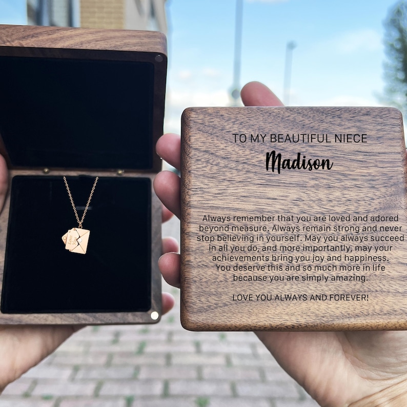 Może przedstawiać: Drewniane pudełko z złotym naszyjnikiem w środku. Pudełko ma wygrawerowany napis: "Do mojej pięknej siostrzenicy Madison. Pamiętaj zawsze, że jesteś kochana i uwielbiana ponad wszelką miarę. Zawsze bądź silna i nigdy nie przestawaj wierzyć w siebie. Niech ci się zawsze udaje we wszystkim, co robisz, a co najważniejsze, niech twoje osiągnięcia przynoszą ci radość i szczęście. Zasługujesz na to i o wiele więcej w życiu, ponieważ jesteś po prostu niesamowita. Kocham cię zawsze i na zawsze!"