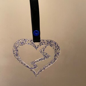 May include: A clear heart-shaped ornament with blue glitter and a lightning bolt design. The ornament is attached to a black leather strap with blue buttons.