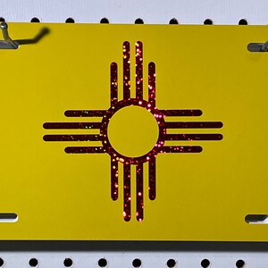 May include: A yellow license plate featuring the Zia symbol of New Mexico. The Zia symbol is a circle with four groups of four rays extending outward, filled with red glitter. The plate has mounting holes.
