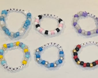 May include: Six colorful beaded bracelets with various names spelled out in white letter beads. The bracelets feature a variety of colors, including blue, pink, purple, yellow, and red. Some bracelets have star-shaped beads.