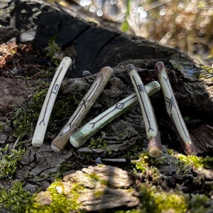 Może przedstawiać: Pięć drewnianych patyczków z wyrzeźbionymi symbolami na porośniętym mchem leśnym podłożu. Patyczki są jasnobrązowe i mają naturalne wykończenie.