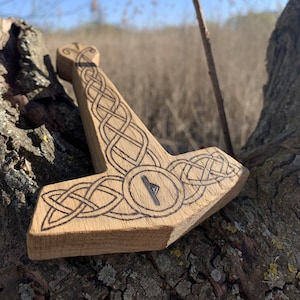 Può includere: Una scultura in legno del martello di Thor, Mjolnir, con intricati nodi e la runa di protezione, Algiz, al centro. Il martello è appoggiato su un tronco d'albero.