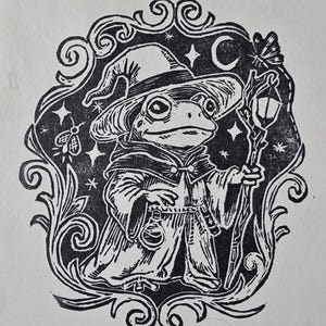 May include: A black and white linocut print featuring a frog dressed in a robe and pointed hat. The frog holds a staff with a lantern, a small bottle hangs from its belt, and it is surrounded by a decorative border with stars and a moth.