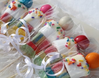May include: A close-up of several individually wrapped candy skewers. Each skewer has a marshmallow topped with white chocolate and sprinkles, gummy candies, and a round candy on top. The skewers are tied with white ribbon.