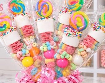 May include: A bouquet of five individually wrapped candy skewers arranged in a pink scalloped container. Each skewer features a colorful swirled lollipop, a large white marshmallow topped with rainbow sprinkles, and various gummy candies.