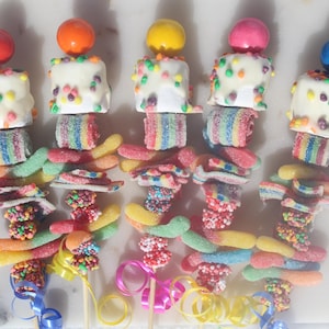 May include: Five colorful candy skewers with a marshmallow topped with white chocolate and sprinkles, rainbow gummy candies, and a round candy on top.