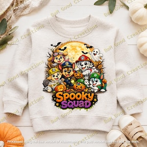 May include: A light heather gray crewneck sweatshirt featuring a colorful graphic of cartoon puppies dressed in Halloween costumes with pumpkins and a ghost against a full moon background. The text Spooky Squad is printed in orange and purple.