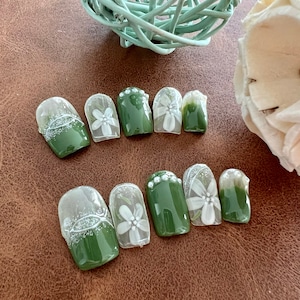 Könnte beinhalten: Zehn grüne und weiße Nagelspitzen mit einem Blumenmuster. Die Nagelspitzen sind in zwei Reihen angeordnet.
