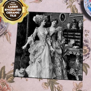 Op de afbeelding: Zwart-witfoto van twee vrouwen in historische kleding. De vrouw links houdt een riem vast die aan een kleine witte hond is bevestigd. De vrouw rechts leest een brief. De foto is omlijst met een zwarte rand.