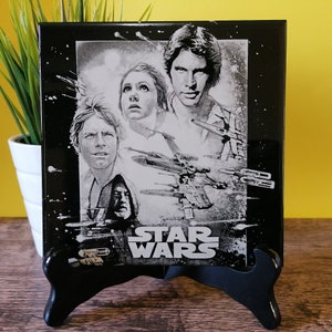 Puede incluir: Azulejo de cerámica en blanco y negro con una imagen de Star Wars con Luke Skywalker, la princesa Leia, Han Solo y Obi-Wan Kenobi. El azulejo tiene un fondo negro con estrellas blancas y el texto "STAR WARS" en blanco.