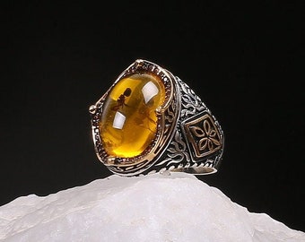 May include: A silver ring with an oval-shaped amber stone set in a gold-toned bezel. The ring band is decorated with intricate floral patterns.