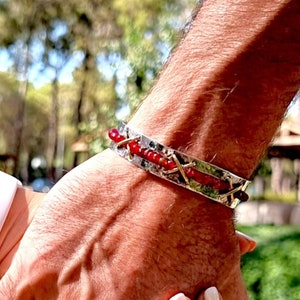 May include: A silver bracelet with red beads and a white smart watch are visible as two people hold hands. The background features a green lawn with yellow flowers and trees.