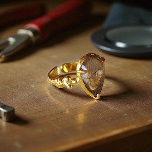 May include: A gold-colored ring with a teardrop-shaped, brown gemstone. The ring's band has a textured, ornate design. The ring is displayed on a wooden surface, with tools and other objects in the background.