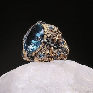 May include: A gold and black ring with a large blue gemstone surrounded by smaller blue gemstones. The ring is set on a white rock.