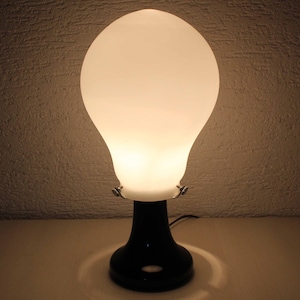 Puede incluir: Una lámpara de mesa de vidrio blanco esmerilado con forma de bombilla y base negra.