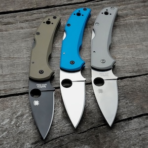 May include: Three folding knives with different colored handles: olive green, bright blue, and gray. Each knife has a silver blade with a curved edge and a round hole near the base. The blades are marked with "Spyderco CPM S30V".