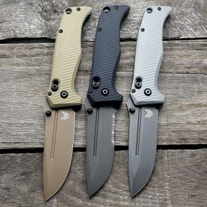 May include: Three folding knives with different colored handles. The knives have a black blade with a serrated edge. The handles are tan, black, and gray.