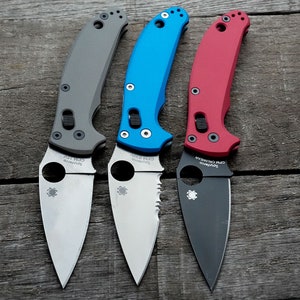 May include: Three Spyderco pocket knives with different colored handles. The first knife has a gray handle and a silver blade. The second knife has a blue handle and a silver blade. The third knife has a red handle and a black blade. All three knives have the Spyderco logo on the blade.