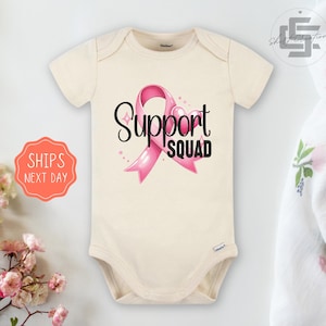 May include: A cream-colored baby bodysuit with a pink ribbon and the text "Support Squad".