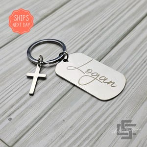 May include: A silver-colored keychain with a cross charm and a rectangular tag engraved with the name "Logan" in cursive. The keychain includes a split ring for attaching keys. The background is a light gray wood grain.