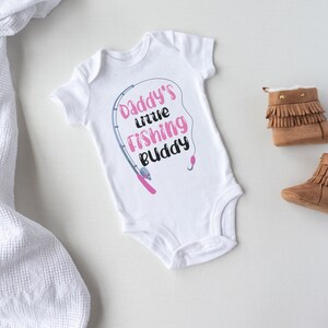 May include: White baby onesie with a pink fishing rod and the text "Daddy's little fishing buddy".