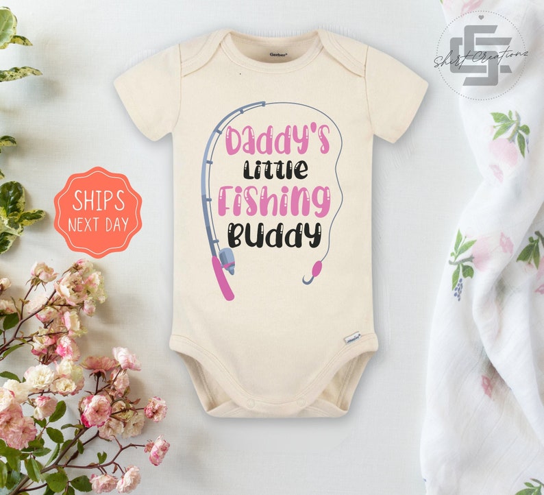 May include: A cream-colored baby onesie with a pink fishing rod and the text "Daddy's little fishing buddy".