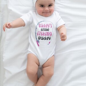 May include: White baby onesie with a pink and gray graphic that says "Daddy's little Fishing Buddy" with a fishing rod and hook.