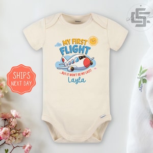 May include: A cream-colored baby bodysuit with a cartoon airplane and the text "My First Flight...But it won't be my last! Layla".