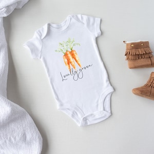 May include: White baby onesie with a watercolor illustration of three carrots and the text "Locally grown." The onesie is paired with brown suede baby boots with fringe.